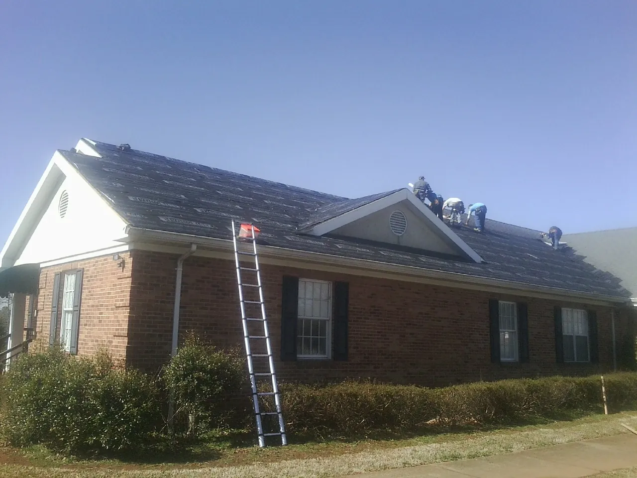 Crew tearing shingles off a brick ranch roof with a ladder set — Emergency Roof Repair work by our crew in Port Richey